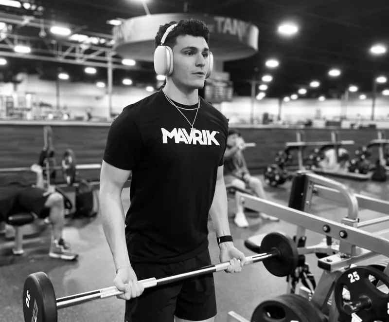 A lifter in a MAVRIK tee carrying a loaded barbell across a gym floor